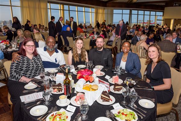 A group of six well-dressed adults pose for a photo at a formal dinner event. They are all seated at the table. The table is set with glasses, plates of cake, salads, rolls, and bottles of wine and water. The background shows a large room filled with other guests seated at tables, with artwork on ea